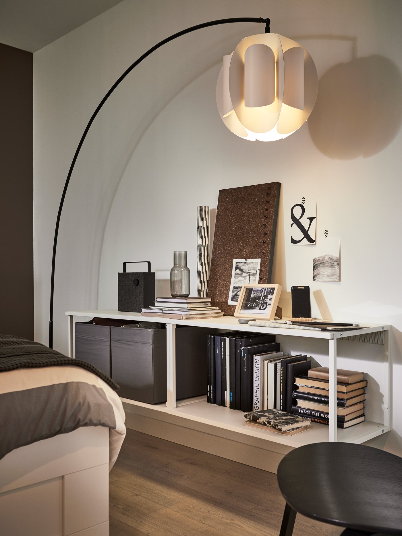 Salón con una estantería abierta blanca, una lámpara de pie de arco con pantalla de lámpara y un tablero de notas marrón.
