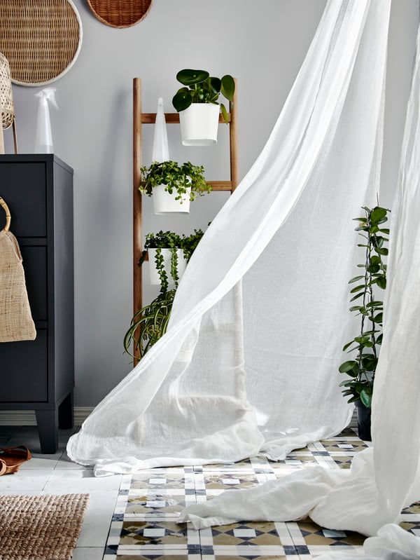 A white curtain is blown into a bedroom by the wind. Behind the curtain is a SATSUMAS plant stand with plants on it.