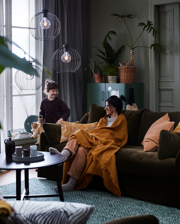A mother sits on the couch under a plaid and smiles at her daughter who is standing next to the couch with a stuffed animal.