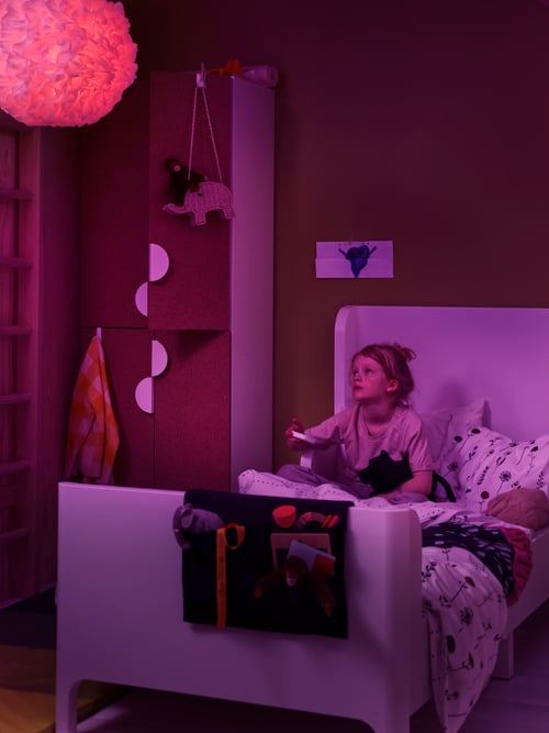 Fetiță în dormitorul copiilor uitându-se la o lampă suspendată și ținând o telecomandă pentru becul colorat cu LED-uri.