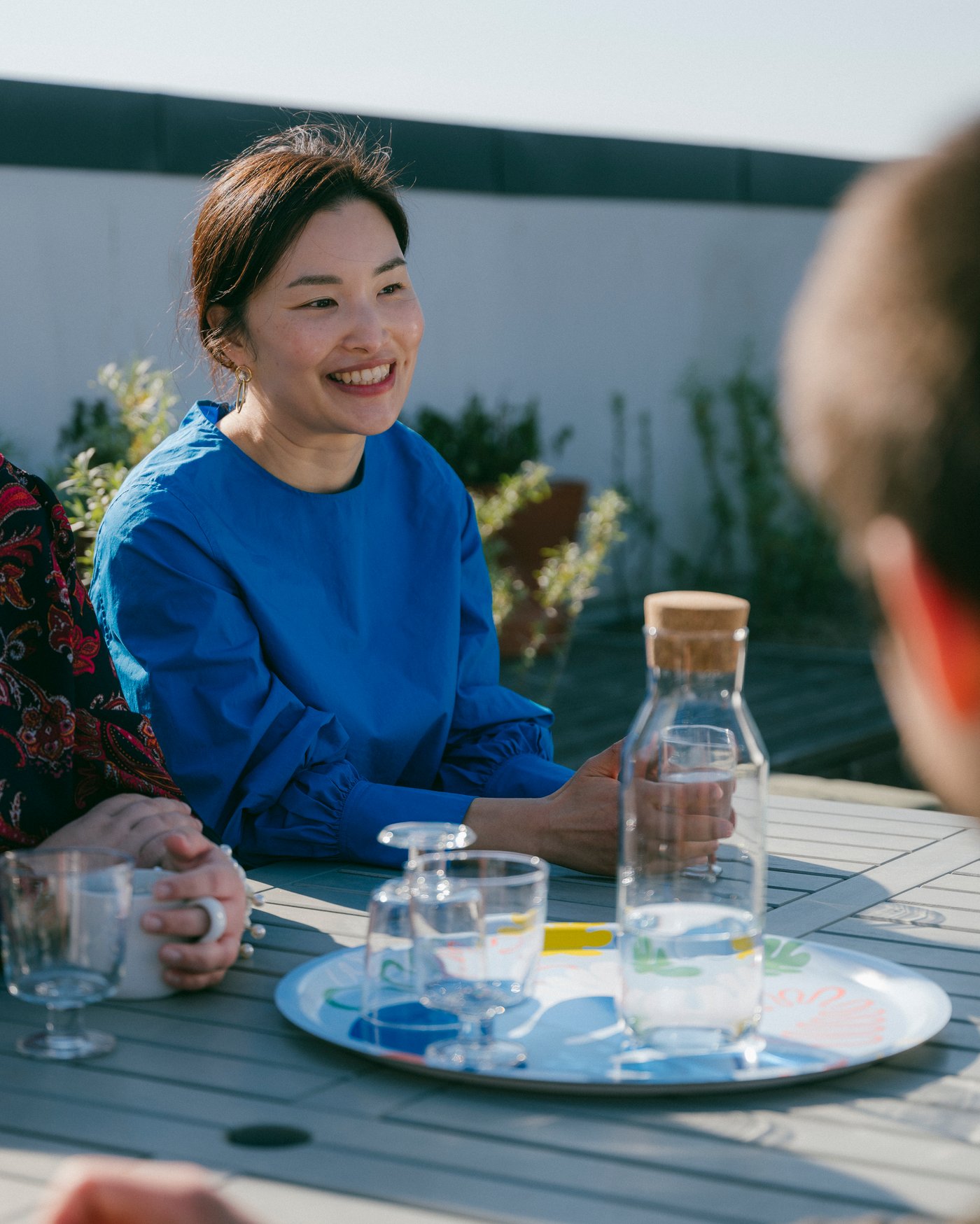 Middle aged woman sitting at outdoor picnic table smiling and others. Small tray sits on table with glasses and large bottle of water
