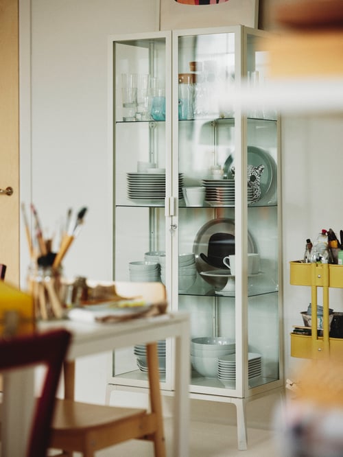 Un dulap de ușă din sticlă MILSBO care conține o carafă IKEA 365+ și mai multe boluri și farfurii se află lângă un cărucior RÅSKOG galben.