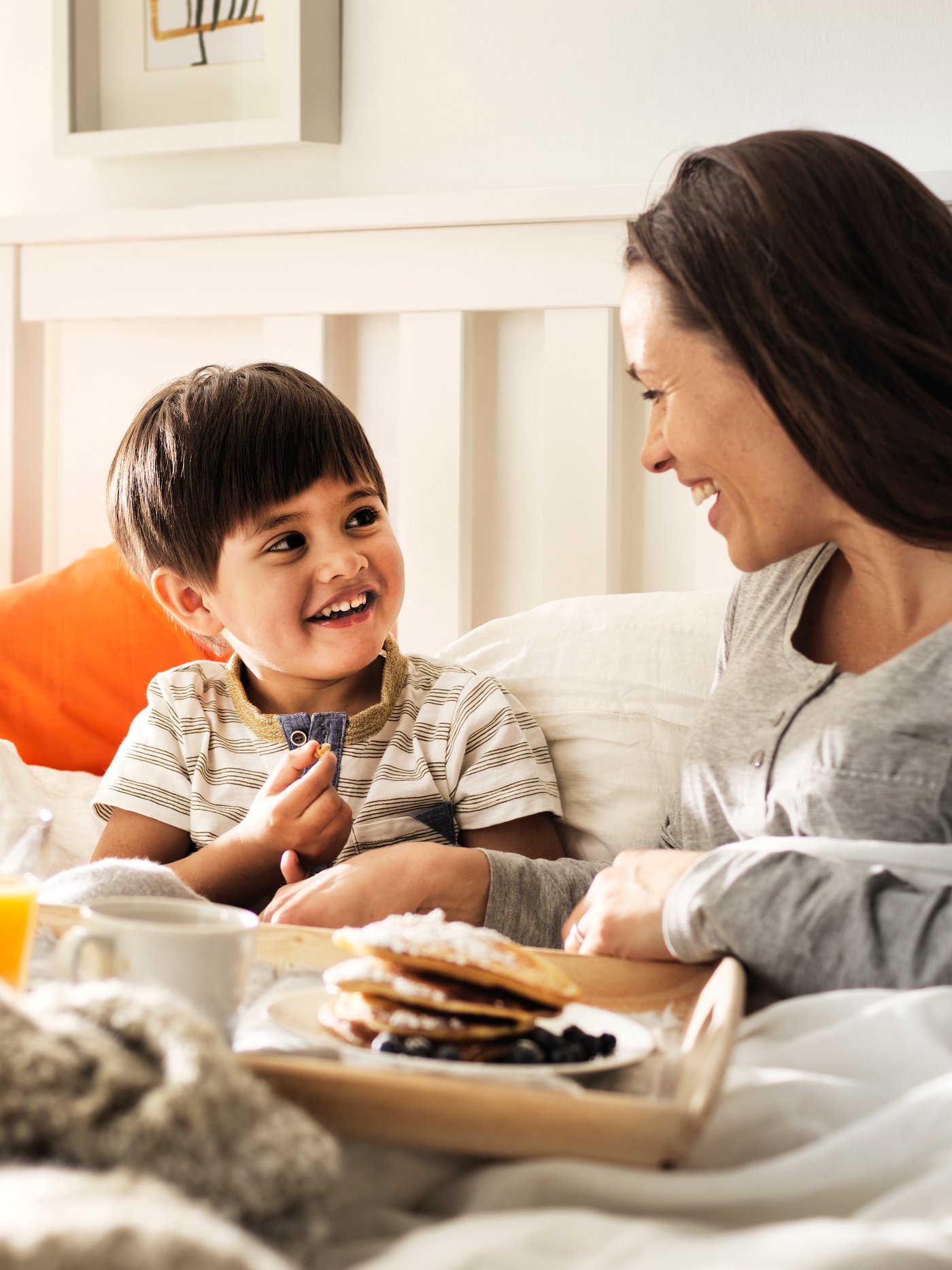 En mor og et barn sidder i en seng med en bakke med morgenmad. 