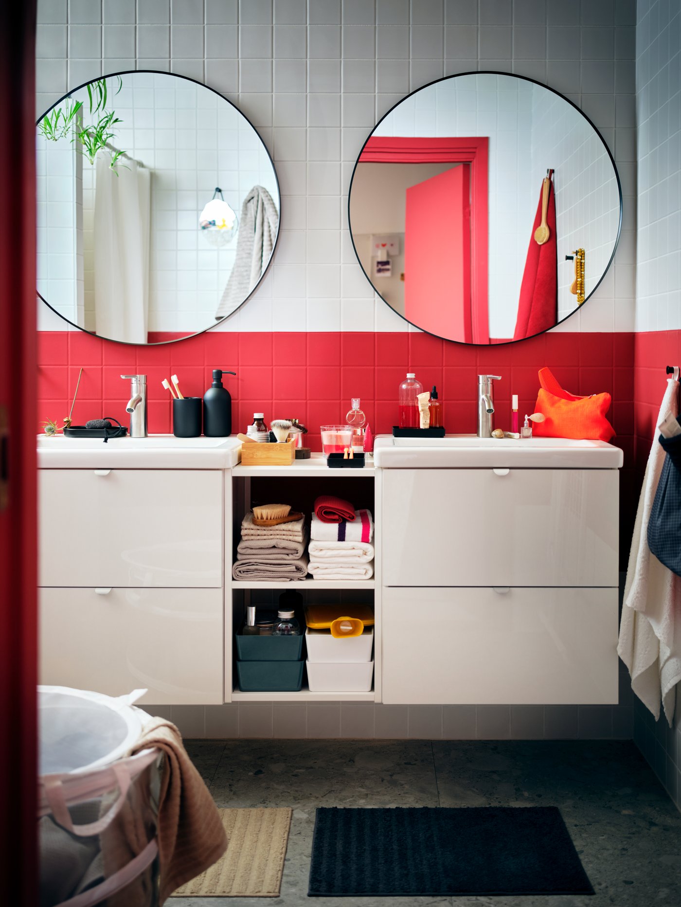 Un baño moderno con dos espejos redondos, un cesto para la ropa redondo, un armario bajo con estantes y dos alfombrillas de baño.