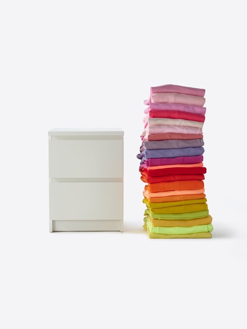 A white MALM chest of 2 drawers set beside a set of stacked colourful fabrics, in a bathroom setting.