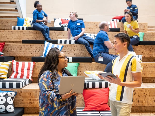 Two IKEA co- workers chatting in the foreground of the photo. Behind them, people in IKEA work clothes are sitting on the stairs.