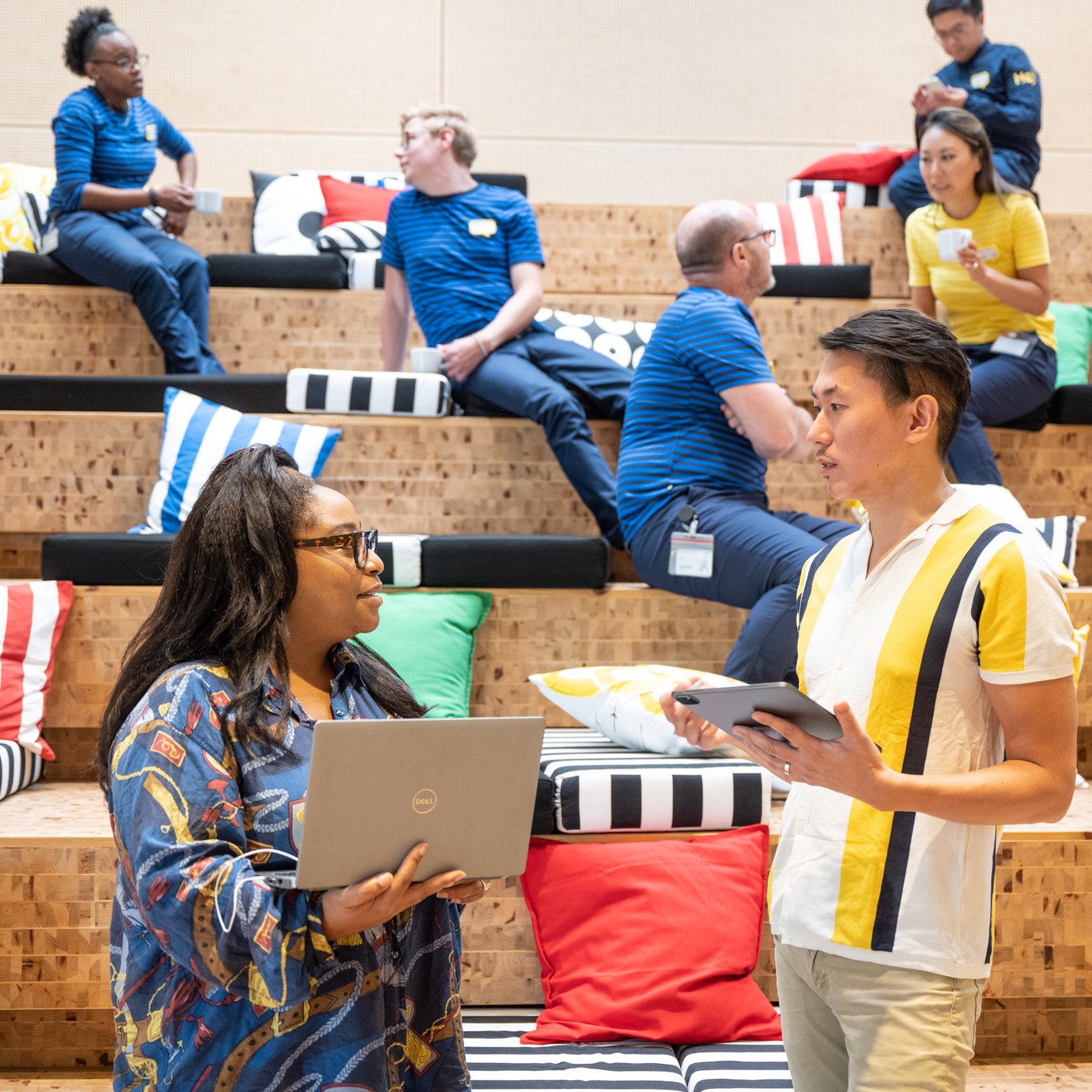 A group of people on wooden stands with colorful cushions, some with laptops or tablets, talking or working.