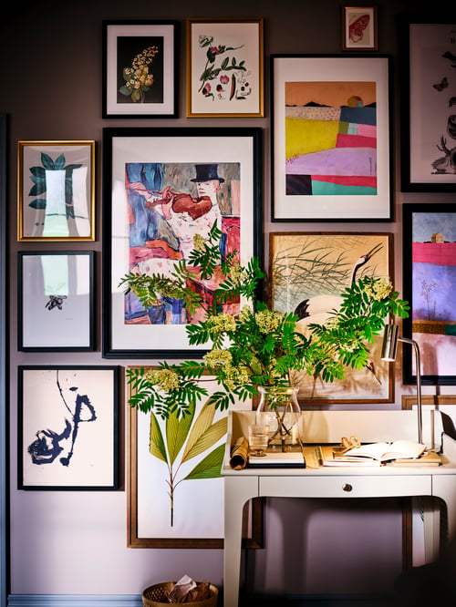 Un vase BEGÄRLIG en verre clair rempli de fleurs sur un bureau LOMMARP, avec des impressions encadrées aux couleurs vives sur le mur derrière.