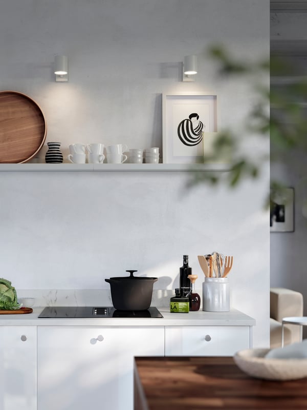 A white MOSSLUNDA picture ledge is placed on a wall above a kitchen stove and it has a picture frame, a tray and cups on it.