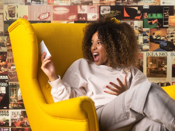 In een gele fauteuil zit een vrouw met krullen in een lichtgekleurde sweater en broek. Ze kijkt verrast naar haar telefoon.