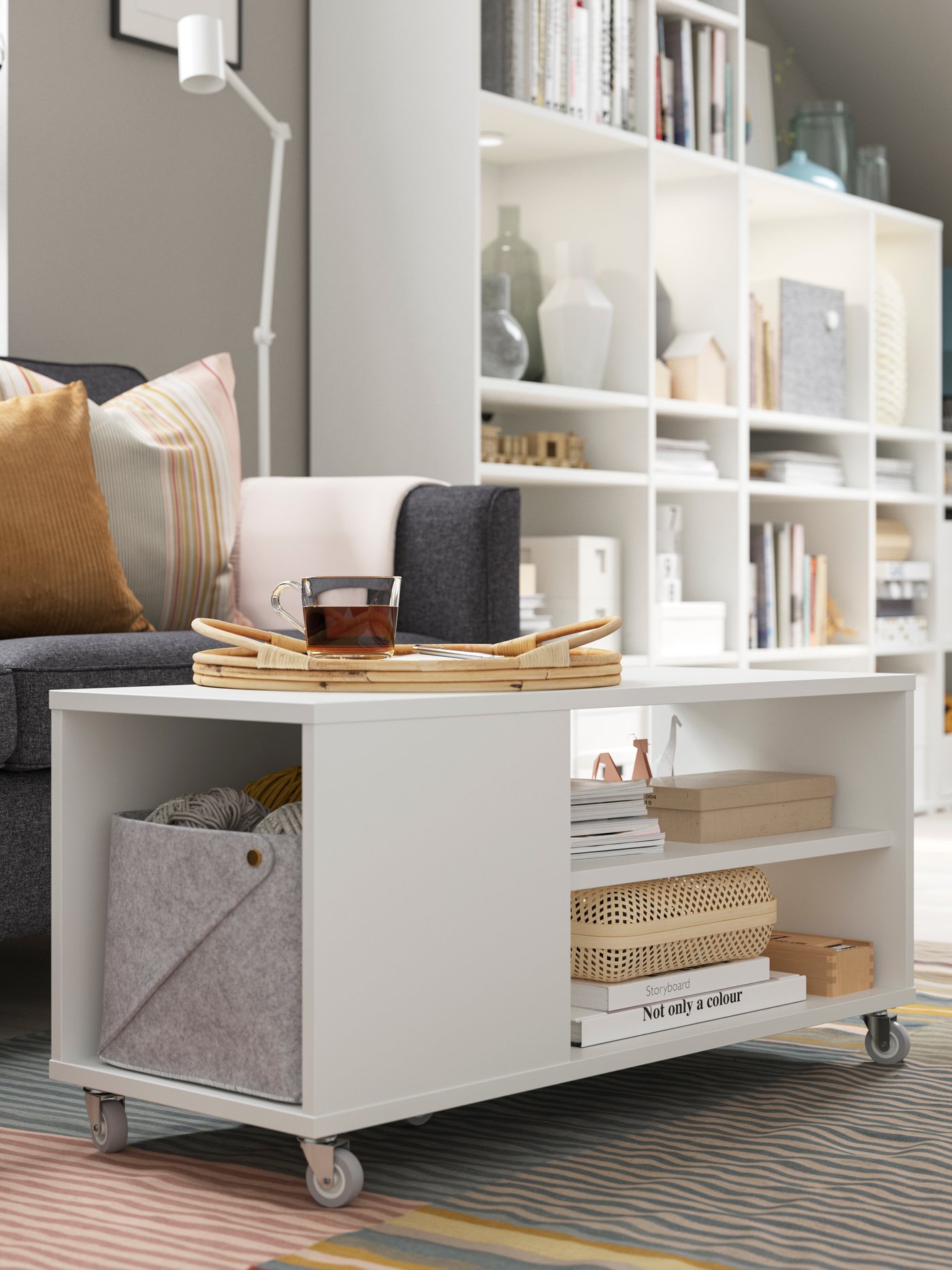 Vit förvaringsmöbel på hjul med öppna hyllor och korg, placerad i ett modernt vardagsrum med bokhylla i bakgrunden.