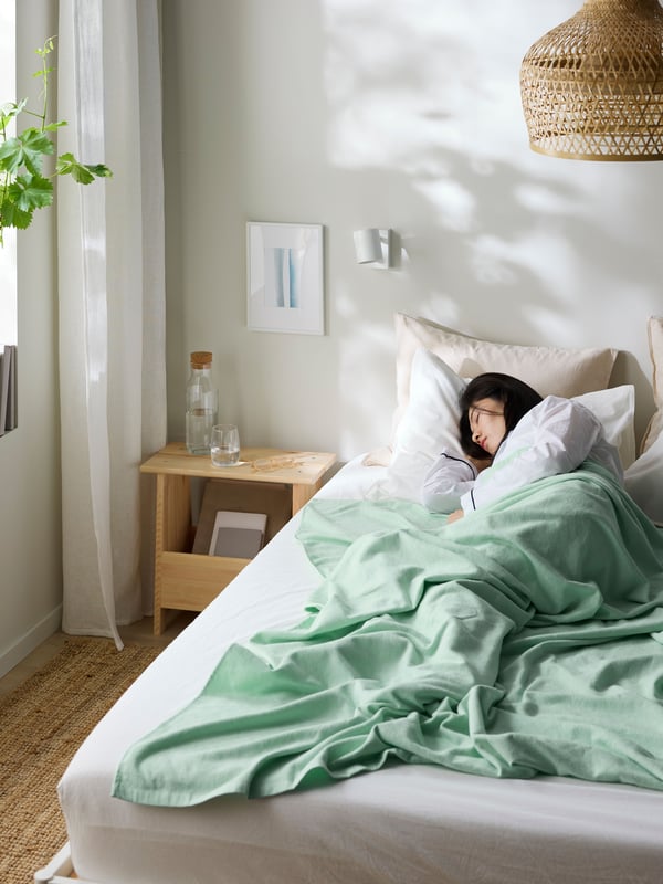 A woman sleeping in bed in an all white/beige bedroom with the light shining through a window at the left.