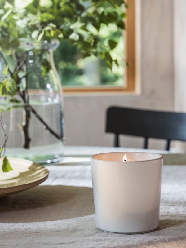 An ADLAD scented candle in a ceramic jar and a clear vase with leafy branches on a dining table close to a window.