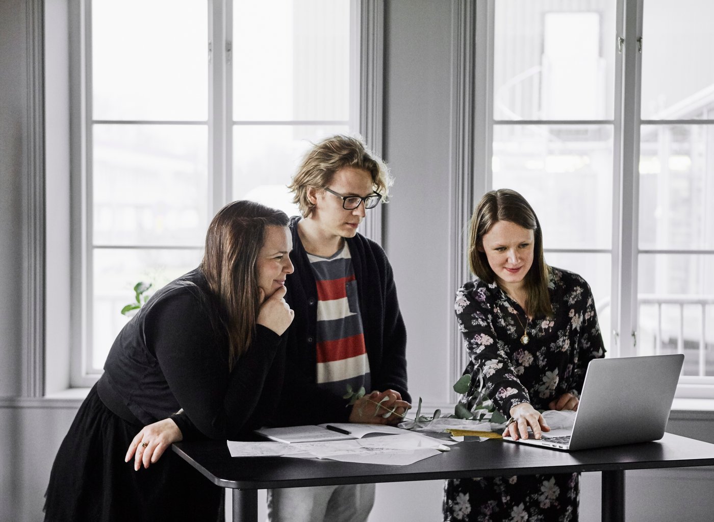 Three people are standing by a table in an office, looking at a laptop.