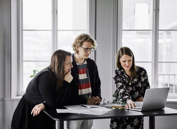 Three people are standing by a table in an office, looking at a laptop.