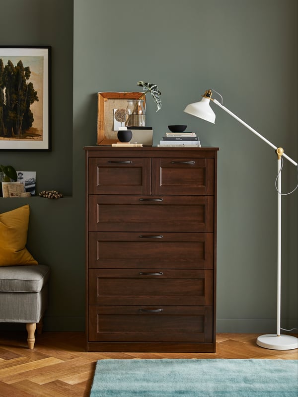On top of white drawers is jewellery in clear storage boxes, a small round mirror and a fancy hat on a mannequin head.