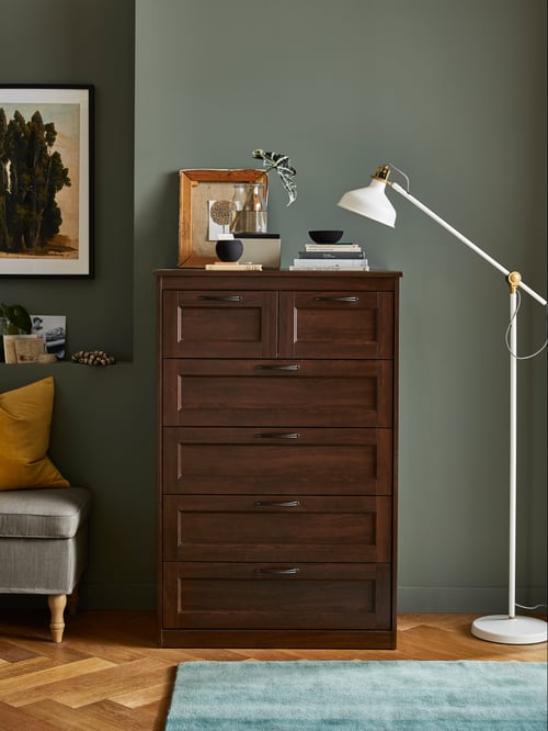 Brown SONGESAND chest of drawers in a dark grey room, next to a painting of trees and a tall white lamp.