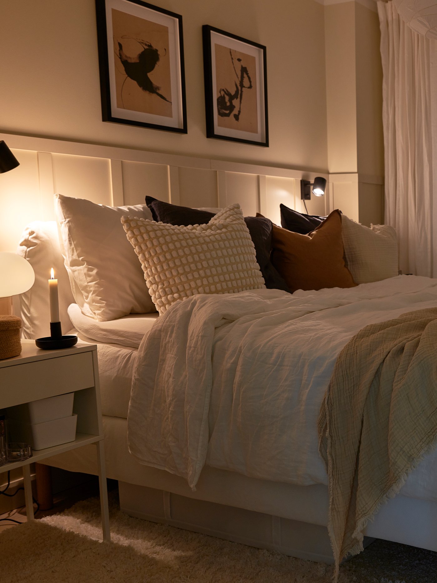 Sovrum med bäddad säng i vitt, kuddar i beige och brunt samt en beige pläd. Sängbord med tändt ljus och två tavlor ovanför sängen.
