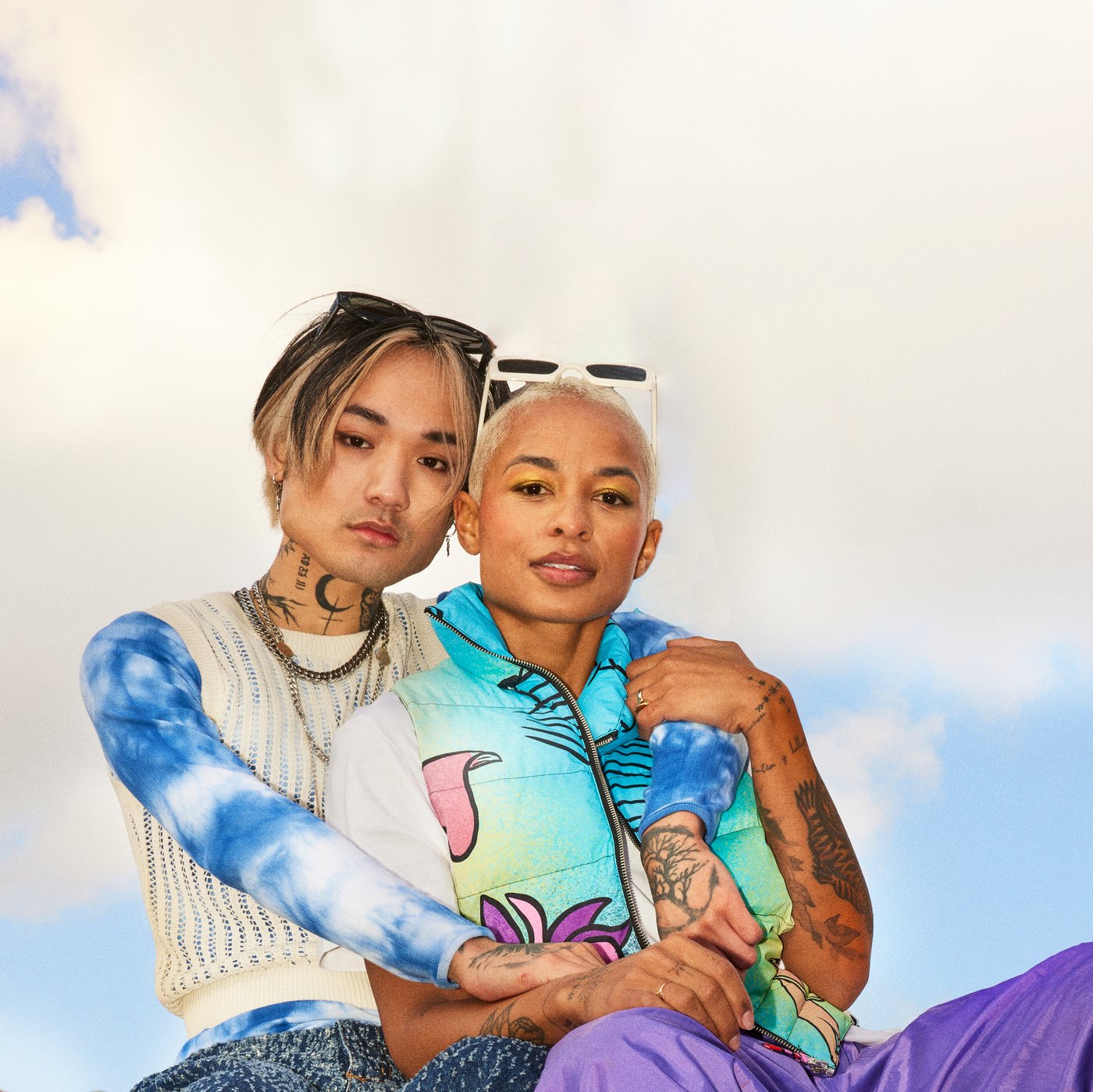 Two people leaning on each other while sitting on a bench outside, a cloudy sky in the background. 