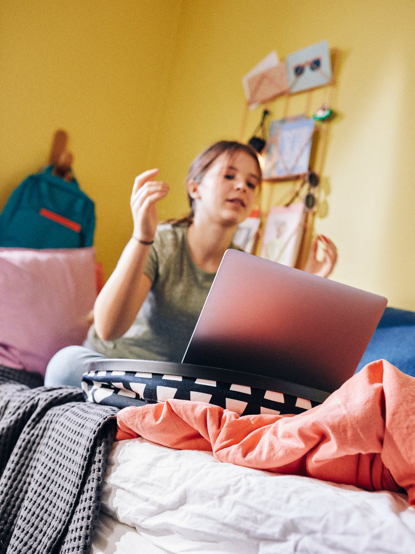 A girl sits on her bed in her room looking at a laptop which is sitting on top of a BYLLAN laptop support.
