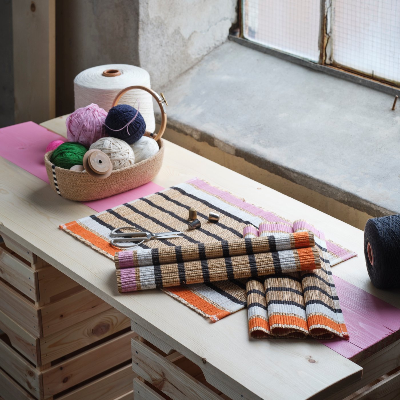 Un cesto di vimini con gomitoli di lana colorati (rosa, verde, bianco, nero) e rocchetti di legno si trova su un tavolo di legno chiaro. Accanto al cesto, ci sono delle strisce di tessuto a righe beige, nere, arancioni e rosa, alcune piegate, e un paio di forbici. Sullo sfondo si intravede una finestra.