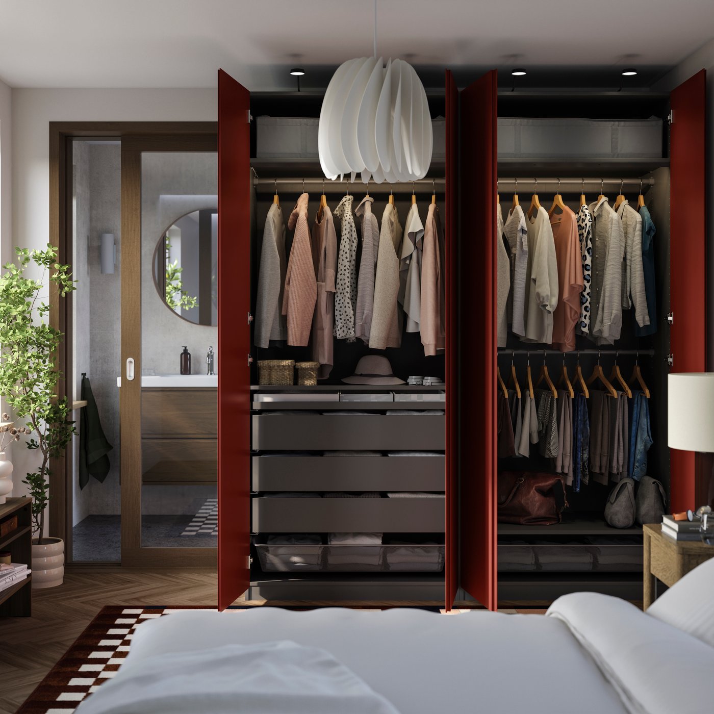 Bedroom with open dark-red wardrobes displaying neatly hung clothes, drawers, handbags, and accessories. A white sculptural pendant light hangs above the wardrobe, with a bed in the foreground and a bathroom visible through a glass door.