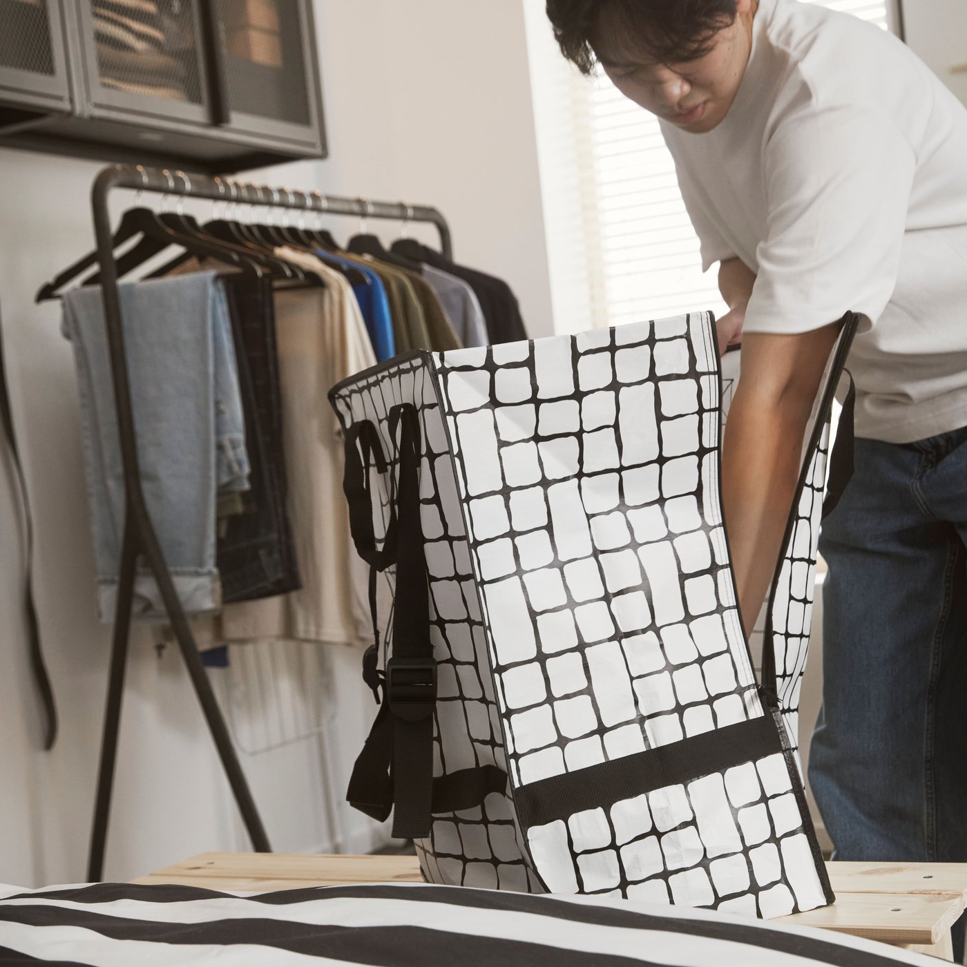 A GÖRSNYGG bag with a black and white grid pattern placed on a bench by a person, with a clothes rack in the background.