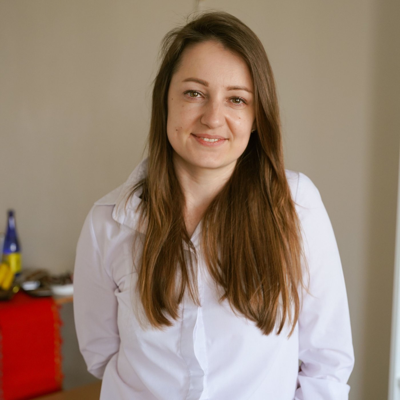 Nataliia v bílé košili s hnědými rozpuštěnými vlasy stojí uvnitř místnosti a dívá se do objektivu fotoaparátu.