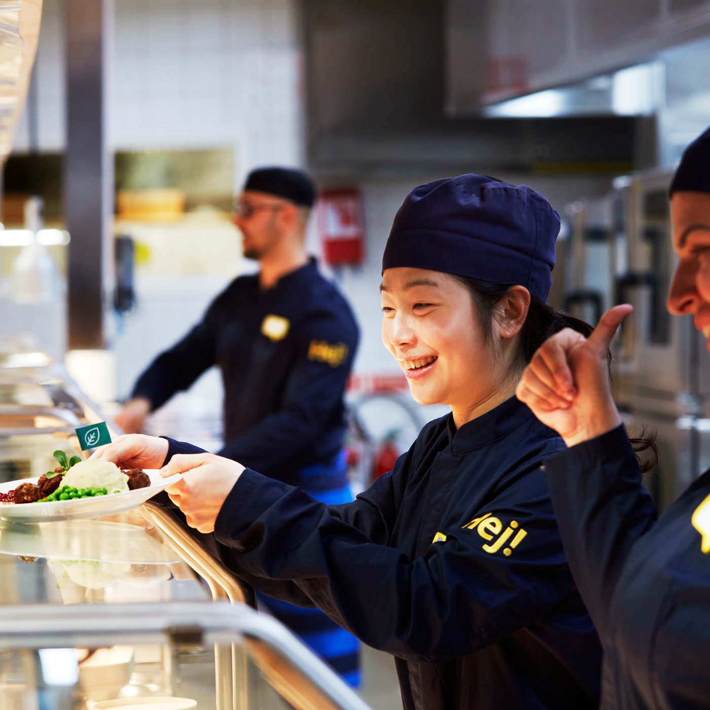 Tre IKEA medarbetare i resturangdisken. Personen i mitten sätter ner en tallrik med plantbullar och potatismos på disken.