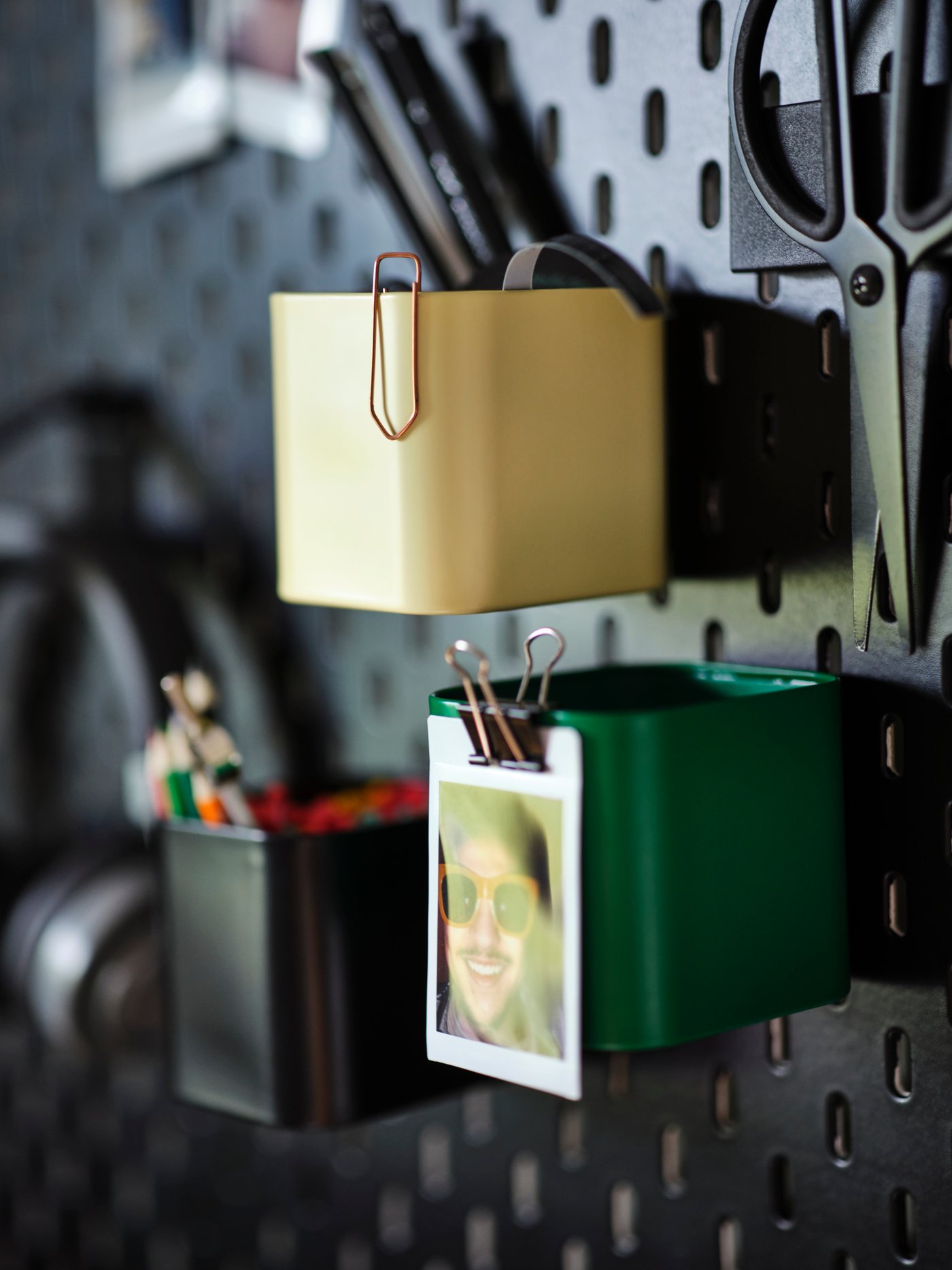 A close-up of an UPPSPEL peg board with multicoloured SKÅDIS containers holding pens, paper clips and a polaroid photograph.