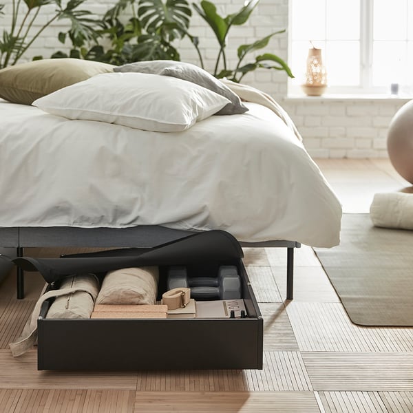 VARDÖ underbed storage with wheels shown opened with stored clothing items under a bedframe with ANGSILILJA bedsheets.