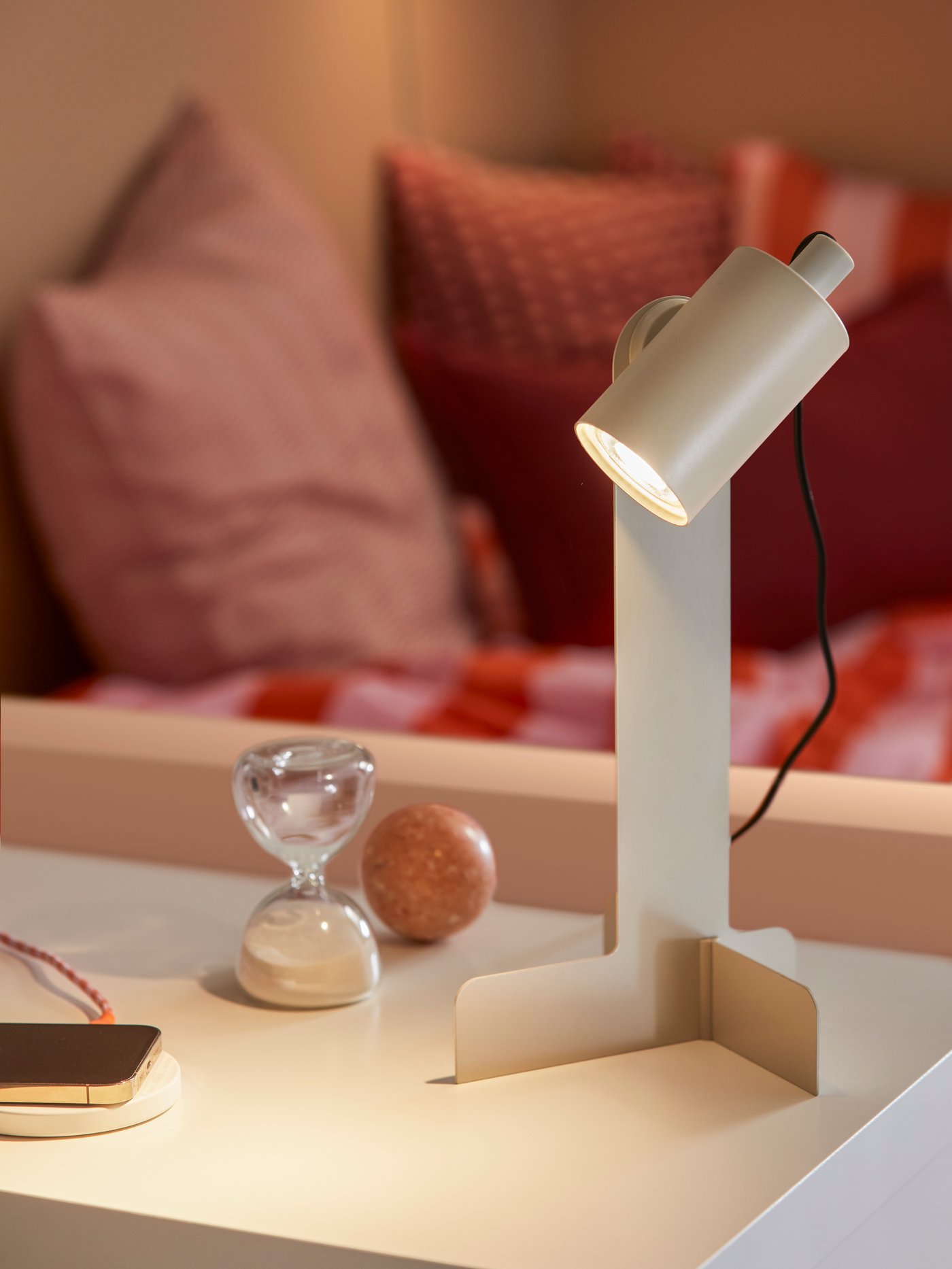 A lit, beige FLOTTILJ desk lamp and a mobile phone on a white wireless charger on a white desk surface by a bed.