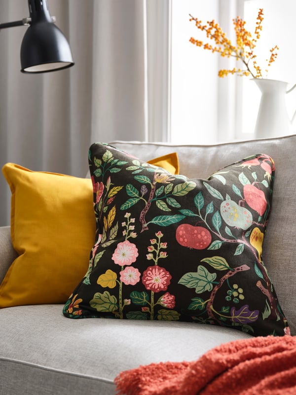 Soffa med färgglada kuddar, inklusive en svart kudde med blommönster och en gul kudde, med ett fönster och en vit vas med orange blommor i bakgrunden.