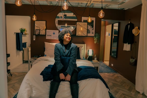 Shadia is seated on a bed, wearing a dark jacket and black boots, with curly hair and a relaxed posture, smiling gently.