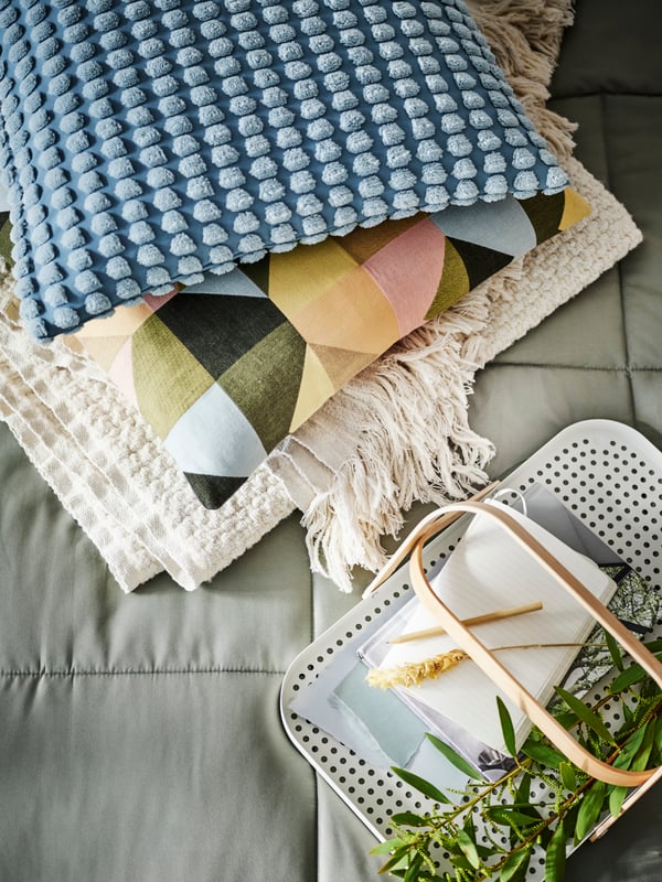 A pile of bed linen on a KÖLAX bedspread includes cushions in a multicolour SVARTHÖ cover and a pale blue SVARTPOPPEL cover. 
