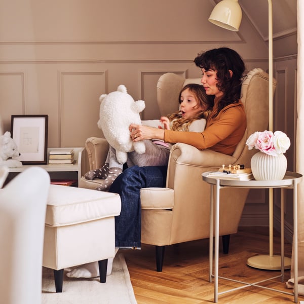 Uma criança está sentada ao colo de um adulto, com um peluche GREJSIMOJS, numa poltrona STRANDMON de abas. Ao lado, encontra-se um pufe STRANDMON.