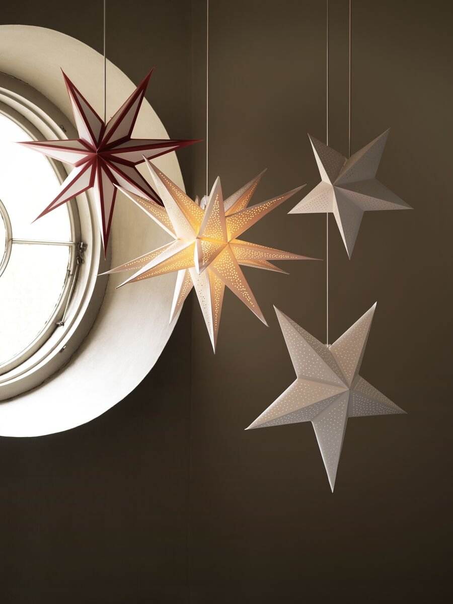 Čtyři hvězdy STRÅLA v různých velikostech visí v malém kulatém okně, přičemž ta prostřední je uvnitř rozsvícená.