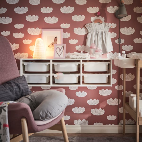 TROFAST wall storage in white with white plastic boxes against a pink wall with a white floral geometric print, with baby items on top and an armchair in front with a grey nursing pillow on it.