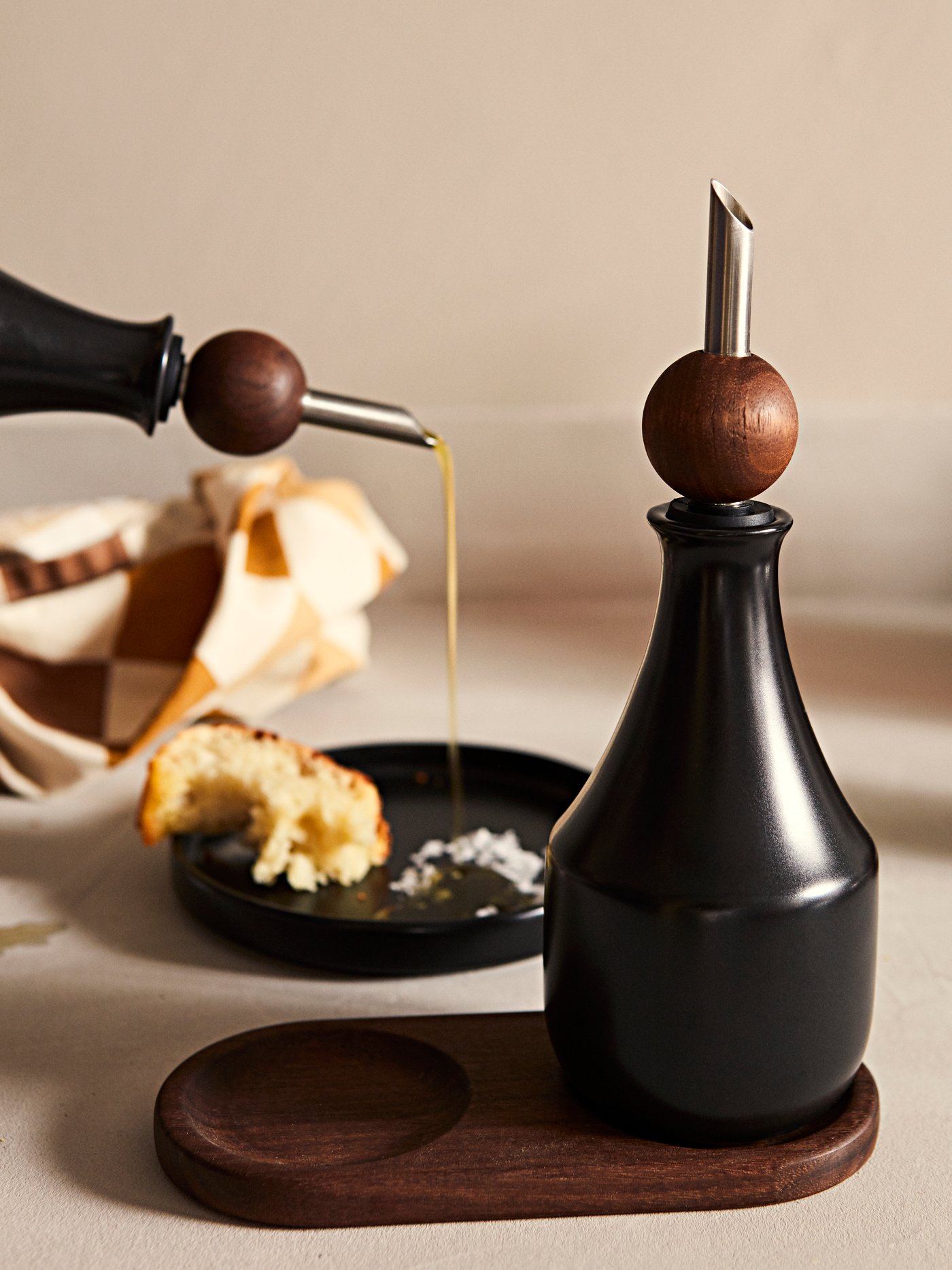 Minyak zaitun sedang dituang ke dalam pinggan kecil KRÖSAMOS yang mempunyai roti dan garam. Satu lagi botol dengan penutup bermuncung KRÖSAMOS terletak di hadapannya.