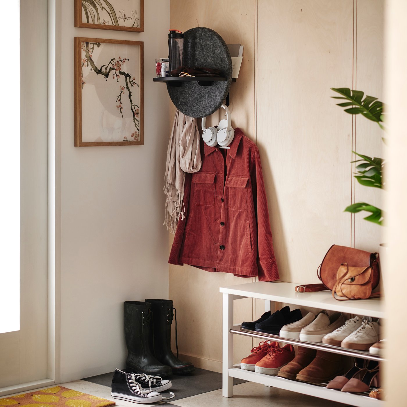 Entré med klädhängare, röd jacka, hatt, skor och väska. Vit skohylla, växt bredvid och två tavlor på väggen.