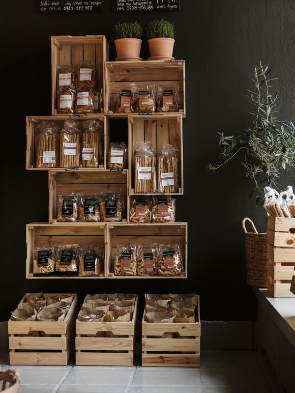 KNAGGLIG trälådor fyllda med matvaror och krukväxter, arrangerade som en väggdisplay i butiksmiljö.