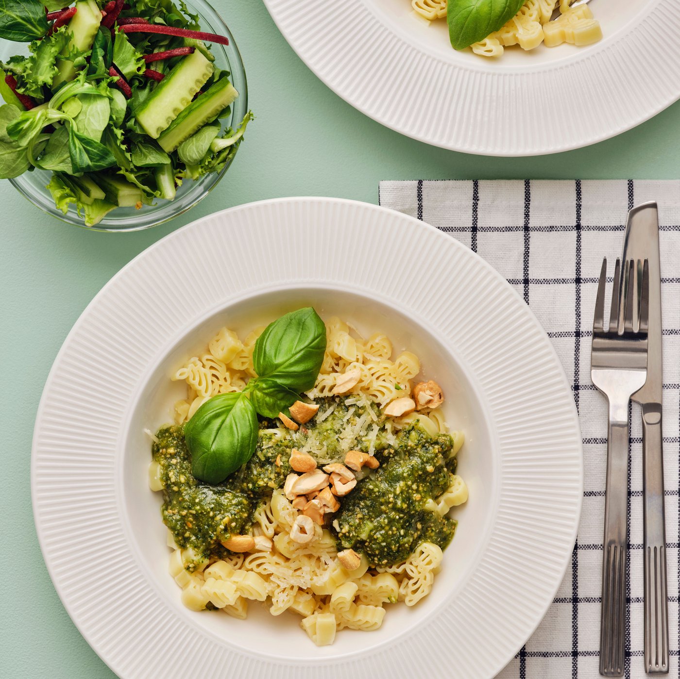 Ein tiefer Teller mit BÄSTISAR Nudeln, grünem Pesto und Cashewnüssen. Daneben liegen ein Messer und eine Gabel.