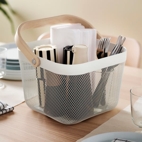 A white RISATORP basket with cutlery, napkins and a salt and pepper mill in it is placed on a wooden dining table.