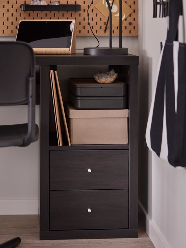 A black-brown KALLAX insert with 2 drawers in a desk with a black swivel chair in the corner of a small home workspace.