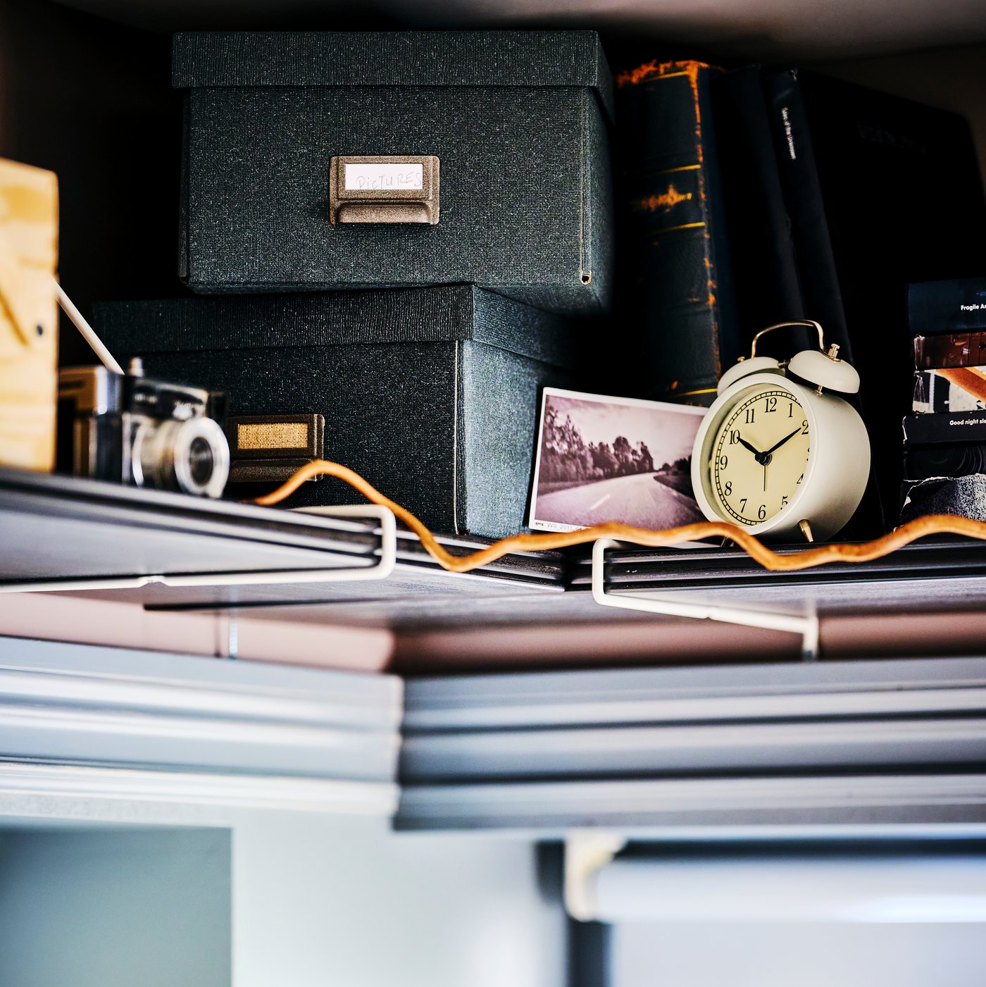 Two TJOG storage boxes with lids on top of one another, with an alarm clock and an analogue camera on a shelf.