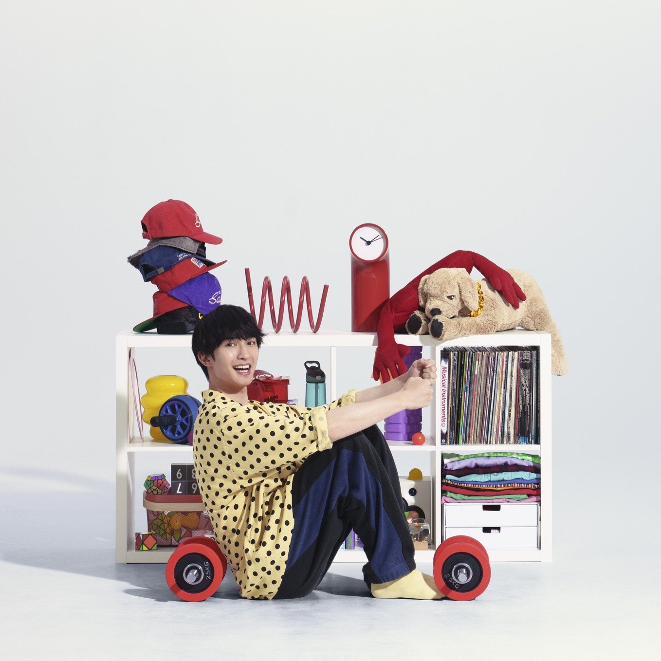  白い収納棚の前に座る人物と、帽子、時計、ぬいぐるみ、レコード、衣類を収めたカラフルな収納。