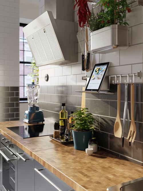 Un comptoir de cuisine a un rail en acier inoxydable KUNGSFORS avec cinq crochets et un support pour tablette. Les ustensiles de cuisine sont accrochés.