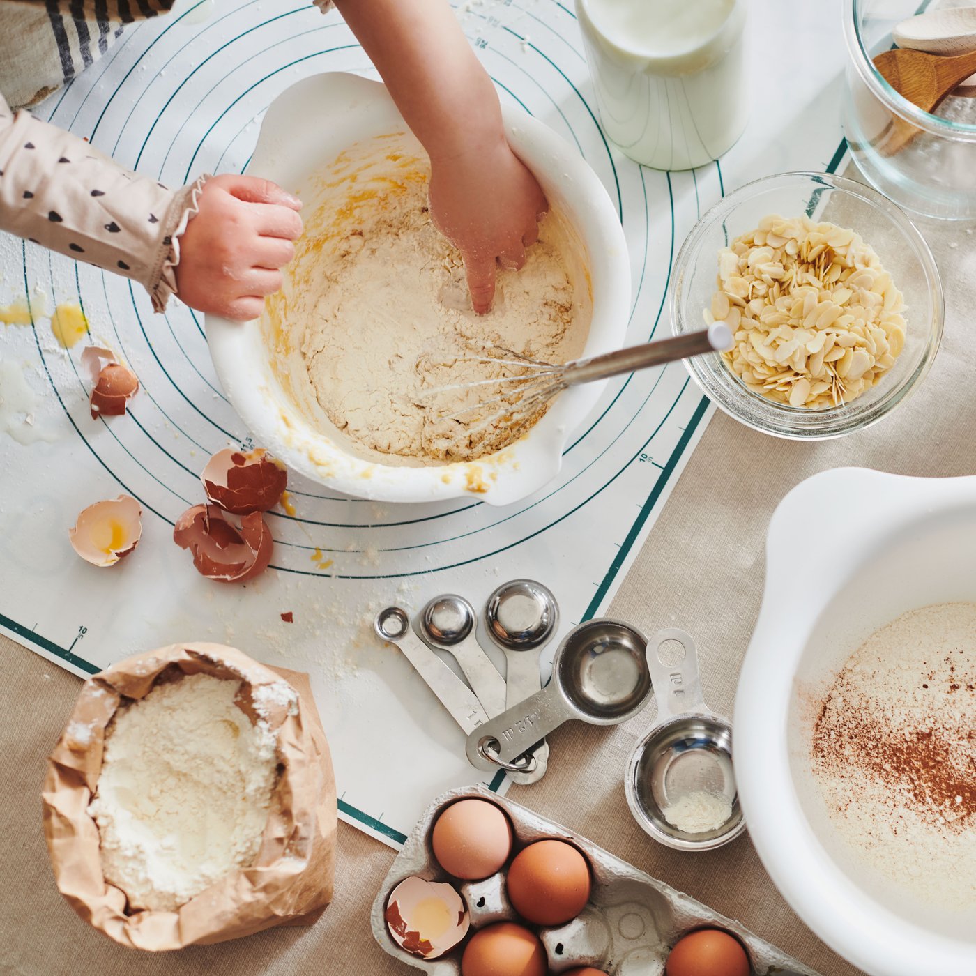 A pair of children’s hands working dough in a white VISPAD mixing bowl on a table with eggs, flour and utensils.