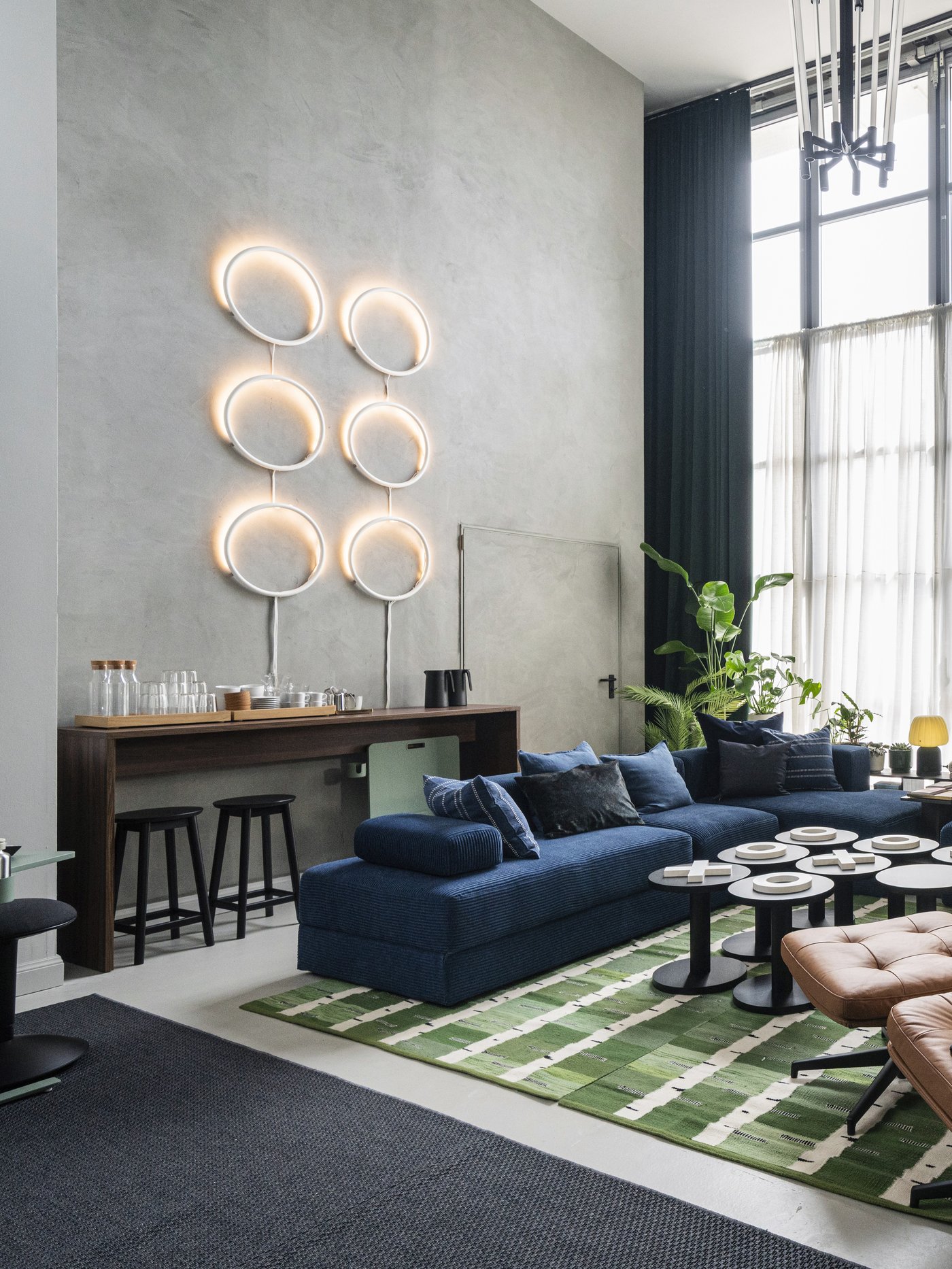 Studio dal soffito alto con sei lampade rotonde sulla parete destra che creano un ambiente accogliente e un divano blu a velluto a coste JÄTTEBO.