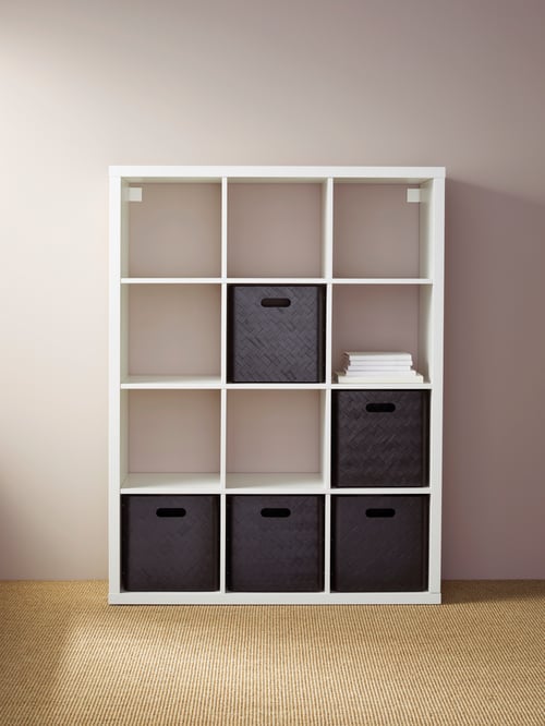 Une étagère KALLAX blanche avec 12 compartiments cubiques est fixée au mur. Elle contient une pile de livres et des boîtes en bambou.